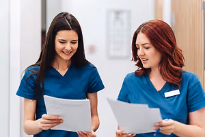 Two women in blue scrubs, standing in a hospital corridor, holding papers and looking at each other with smiles