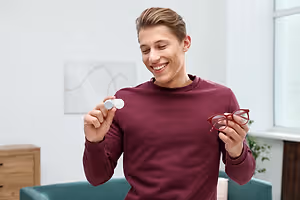 A man is showing a pair of glasses and a contact lens while smiling