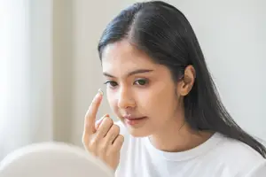 Asian woman wearing a white shirt is putting a contact lens into her eye.