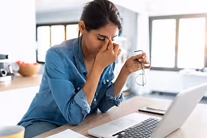 A woman holding glasses and sitting on a chair with her hands covering her face and eyes closed in a kitchen