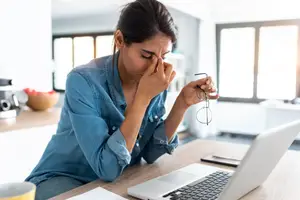 A woman holding glasses and sitting on a chair with her hands covering her face and eyes closed in a kitchen