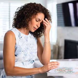 A woman with curly hair is reading a book and holding her head in her hand, probably in the living room.