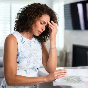 A woman with curly hair is reading a book and holding her head in her hand, probably in the living room.