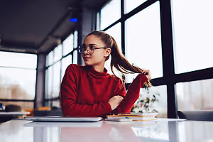 A girl wearing glasses sits at a table with a laptop and books in front of her
