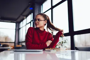 A girl wearing glasses sits at a table with a laptop and books in front of her