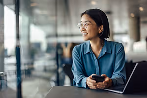 A woman wearing glasses is sitting on a chair while smiling and looking at her phone, with a laptop on a table and a glass wall behind her. In the blurry background, there are people sitting on chairs and a glass wall, with lights on the ceiling and reflections on the glass wall.