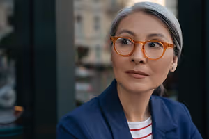 A woman with gray hair wearing glasses and a blue blazer standing and looking to the left in a city street scene