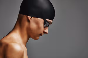 A man wearing a black swim cap and goggles is looking down with a serious expression on a gray background
