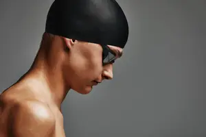 A man wearing a black swim cap and goggles is looking down with a serious expression on a gray background