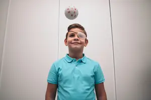 A young boy wearing a blue shirt and eye patch is smiling and looking up at a ball that is floating in the air