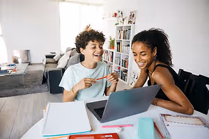 A young boy and his mother are sitting at a desk with a laptop, smiling and holding a marker pen, surrounded by books, papers, and a potted plant on a shelf in the background.