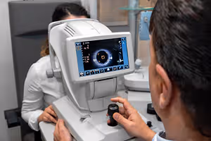 An optometrist is examining a patient's eye using a machine with a screen and buttons in a clinical setting.