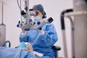 A female ophthalmologist is using a microscope to examine a patient's eye in a medical setting.