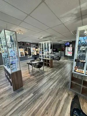 An indoor area with a desk, a chair, a monitor, and a glass cabinet with several items inside, a wooden floor, a gray wall, a ceiling with lights, and a door at the back.