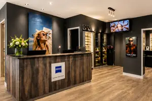 Interior of an optician shop featuring a wooden reception desk, a large poster of a woman wearing sunglasses, a television, and a sign for Zeiss on the desk.
