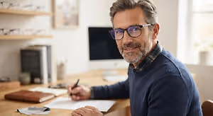 An older man with a beard wearing glasses and a sweater sitting at a desk writing