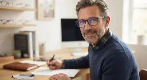 An older man with a beard wearing glasses and a sweater sitting at a desk writing
