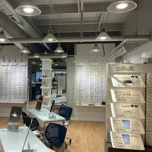 An interior of an optical shop with various items like chairs, tables, and lamps. A desk with a monitor, a bottle, and a lamp is on the left. A cabinet with shelves and glasses is on the right. A glass wall with the word Silhouette on it is behind the cabinet. A glass wall with a tag is on the left.