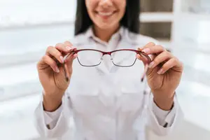 A woman wearing a white lab coat is holding a pair of glasses with a red frame and clear lenses