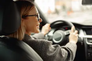 A woman is driving a car while smiling and holding the steering wheel with her right hand. She is wearing glasses and a gray sweater. The interior of the car has a black steering wheel, a silver gear shift, and a black dashboard. The woman is probably driving on the road with blurry surroundings. The car is parked on the side of the road.
