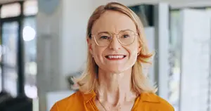 A smiling blonde woman in a bright orange shirt and glasses in an office