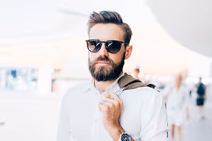 A man with a beard wearing a white shirt and black sunglasses is standing in an outdoor area.