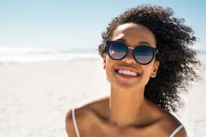 A woman with curly hair smiling and wearing sunglasses on the beach
