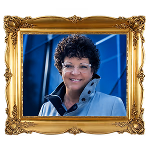 Portrait of a woman with curly hair and glasses wearing a blue jacket and smiling at the camera inside a gold-colored ornate picture frame