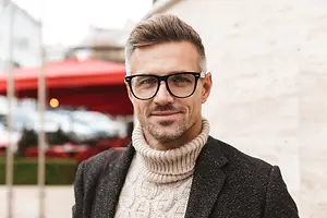 A man with a beard wearing glasses and a black jacket is smiling and looking at the camera