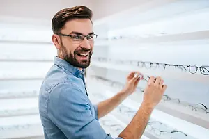 A man wearing glasses is looking at a selection of glasses on a shelf in a shop.
