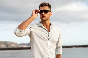 A man in sunglasses adjusts them on his face by a lake.