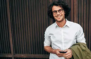 A smiling man with curly hair wearing glasses and a white shirt is standing outside and looking at his phone
