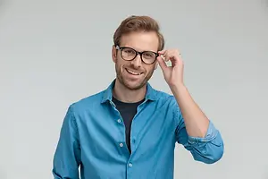 A man wearing a blue shirt and black glasses is smiling and touching his glasses with his right hand.