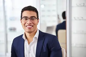 A smiling young man in glasses and a suit is sitting in an office with a glass wall in the background