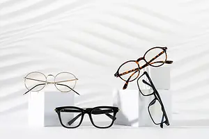 Three pairs of eyeglasses are displayed on a white background with a wavy pattern. The glasses have different frames and lenses, such as round, rectangular, and tortoiseshell. They are placed on top of white boxes, which are arranged in a row. The image has a clean and modern look.