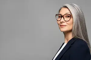 A smiling woman with gray hair and glasses posing for a photo in a gray background