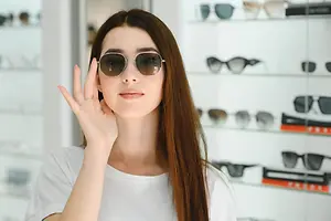A woman with long hair and sunglasses is inside a store, probably taking a selfie.