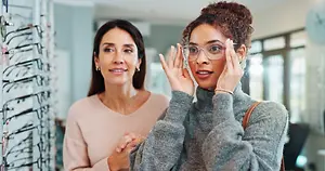Two women in a shop are looking at each other while one of them tries on glasses.