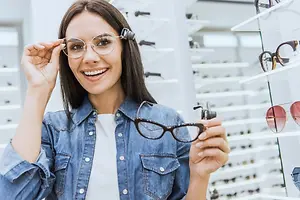 A woman is trying on a pair of glasses in front of a shelf full of glasses in a store.