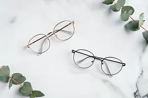 Two pairs of eyeglasses with green leaves on a white marble surface.