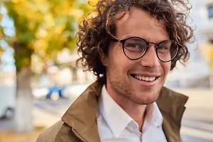 A man with curly hair wearing a jacket and glasses smiles for the camera on a sunny day.