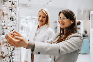 Two women looking at frames in a store, one wearing glasses and a white coat, the other with a gray coat and a bracelet.