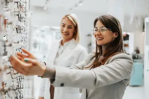 Two women looking at frames in a store, one wearing glasses and a white coat, the other with a gray coat and a bracelet.