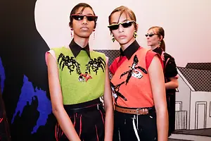 Three women are standing close to each other, probably in a fashion show. They are all wearing sunglasses, a belt, and a short-sleeved top. The woman on the left has a necklace with an earring. The woman in the middle is wearing a shirt with a spider-like print. The woman on the right is wearing a shirt with a bird-like print. Behind them is a white wall with a black and blue design.