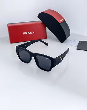 A black Prada sunglasses in a black case with red linings, a red Prada box, and a Prada brochure are displayed on a white surface.