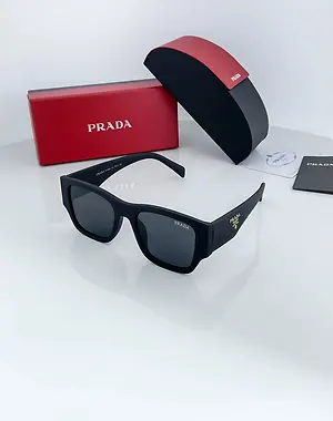 A black Prada sunglasses in a black case with red linings, a red Prada box, and a Prada brochure are displayed on a white surface.