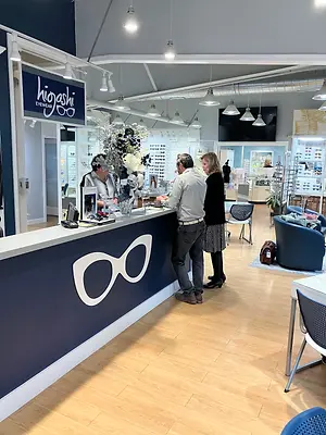 A couple stands at the counter of an eyeglass shop called Higashi, they are talking to the woman at the counter, the shop has a blue theme with wooden floors, and there are chairs and tables around.
