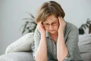 Middle-aged woman in a gray shirt sitting on a couch, wearing glasses and holding her head in pain, probably suffering from a headache