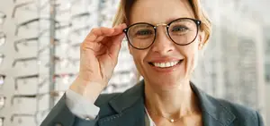 A woman is smiling while adjusting her glasses and holding a pair of glasses in her hand. She is wearing a necklace and a blazer. Behind her, there is a glass wall with many pairs of glasses hanging on it, probably in a store.