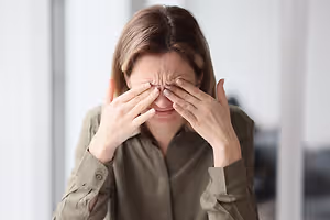 A woman wearing a brown shirt with her hands on her face in front of a window